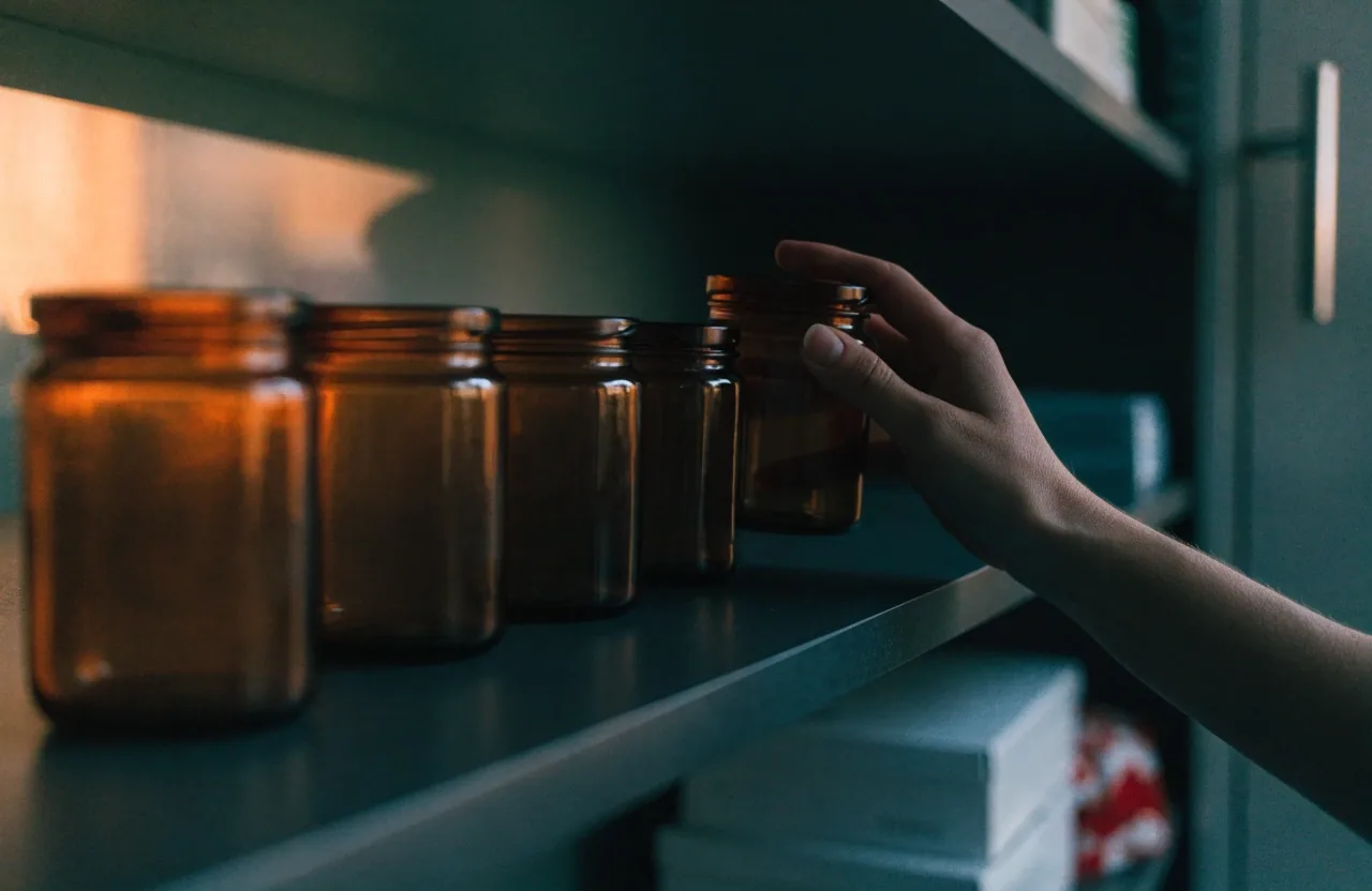 A kitchen shelf at dusk with unlabelled amber jars and a hand lifting one forward for a routine review